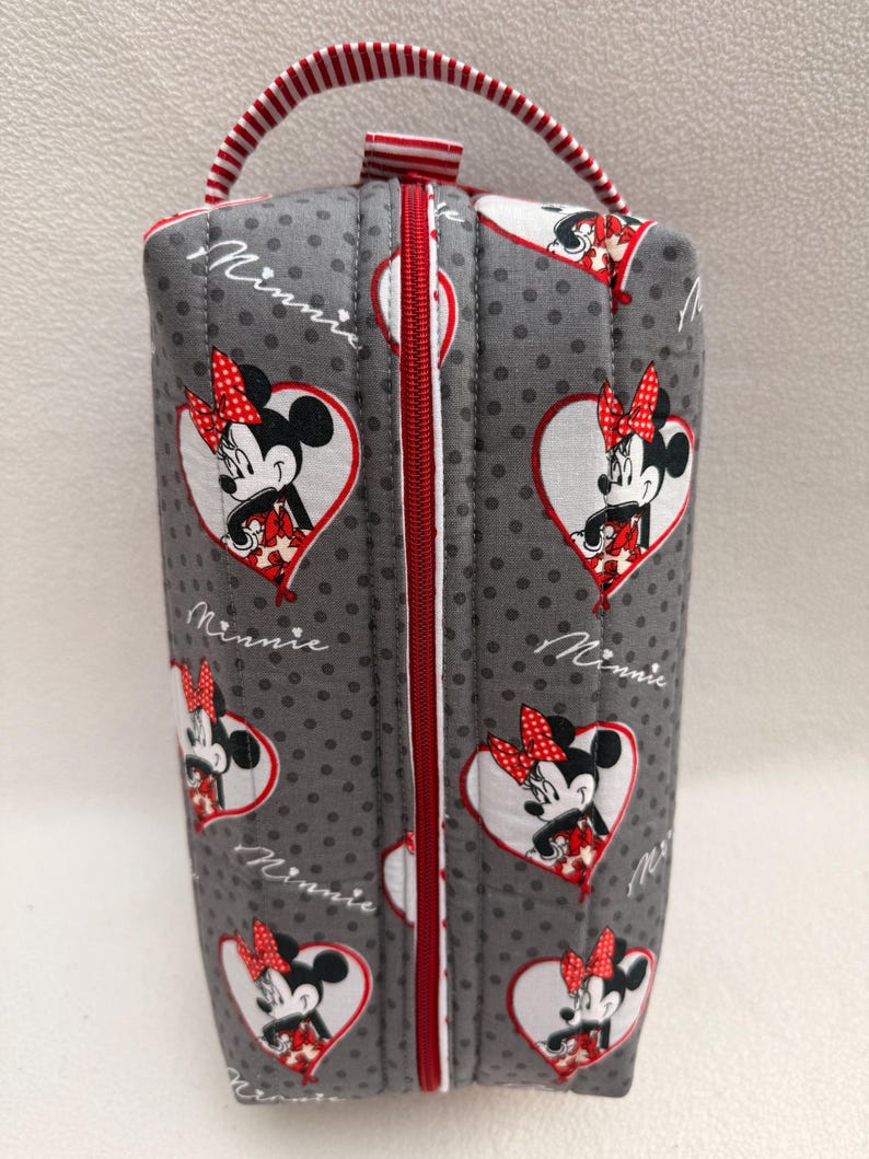May include: A gray, quilted cosmetic bag featuring a pattern of Minnie Mouse in heart-shaped designs. The bag has a red zipper, a red and white striped handle, and the word "Minnie" in script.