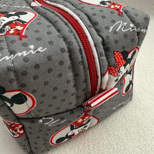 May include: Gray, quilted cosmetic bag featuring a repeating pattern of Minnie Mouse in red, white, and black. The bag has a red zipper with a white border and a small red and white striped fabric loop.