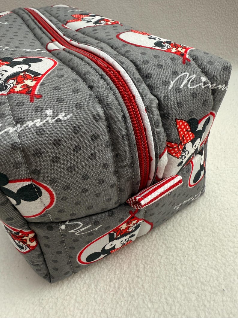 May include: Gray, quilted cosmetic bag featuring a repeating pattern of Minnie Mouse in red, white, and black. The bag has a red zipper with a white border and a small red and white striped fabric loop.