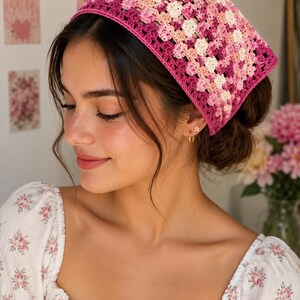 Peut inclure: Un foulard triangulaire en crochet présentant un motif de carrés granny dans des tons variés de rose, magenta et crème. Le foulard possède une bordure unie magenta sur les bords et est réalisé avec un point de tricot texturé et ajouré.