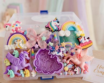 Op de afbeelding: Een doorzichtige plastic opbergdoos gevuld met knutselspullen, waaronder kleurrijke eenhoornfiguurtjes, regenboogbedels, een roze vlinder, een paarse eenhoornvormige koekjesvorm en kleine kraaltjes, met een kaartje met de tekst Unicorn Craft Kit.