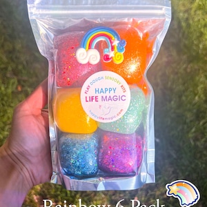 May include: A clear plastic pouch containing six individual portions of colourful play dough in red, orange, yellow, green, blue, and purple, each with glitter. A rainbow charm sits on top, and a circular label reads PLAY DOUGH SENSORY KITS HAPPY LIFE MAGIC.