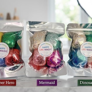 May include: Three silver foil pouches containing colourful glittery play dough balls. Each pouch features a circular white label with the text Happy Life Magic and Play Dough Sensory Kits. The sets are labelled Super Hero, Mermaid, and Dinosaur.