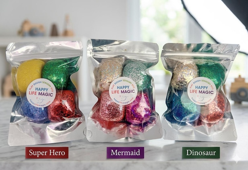 May include: Three silver foil pouches containing colourful glittery play dough balls. Each pouch features a circular white label with the text Happy Life Magic and Play Dough Sensory Kits. The sets are labelled Super Hero, Mermaid, and Dinosaur.