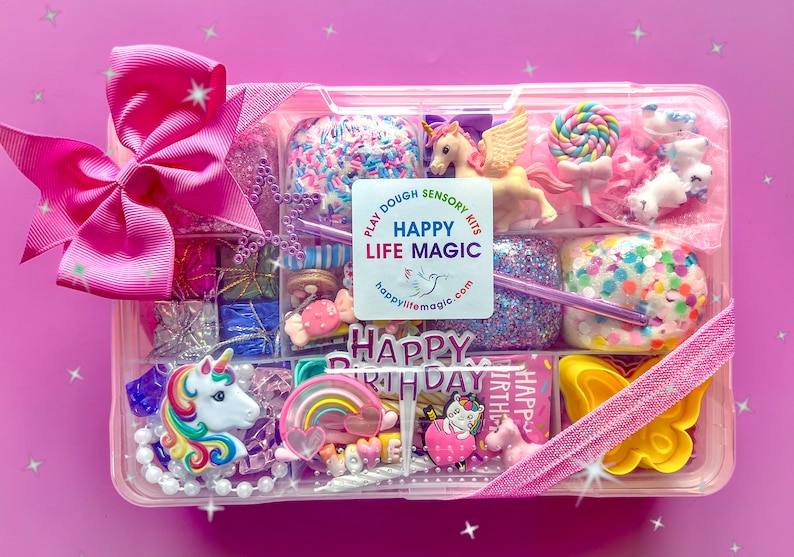 May include: A clear plastic storage container filled with colorful sensory play items, including a unicorn figurine, a rainbow charm, a lollipop, and various beads. A large pink bow is attached to the side, and a label reads HAPPY LIFE MAGIC.