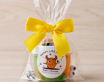 Pode incluir: Um saco de plástico transparente contendo um kit de massa de modelar sensorial, atado com uma fita de cetim amarelo brilhante. O recipiente apresenta um rótulo circular branco com uma ilustração de monstro laranja e o texto Happy Life, Make a Monster Sensory Dough Play Kit.