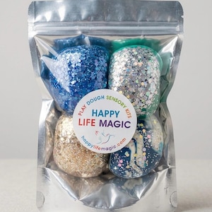 May include: A clear, resealable pouch containing four spheres of glittery play dough in shades of blue, silver, gold, and a marbled navy and white. A circular label on the front reads: Play Dough Sensory Kits, Happy Life Magic, happylifemagic.com.