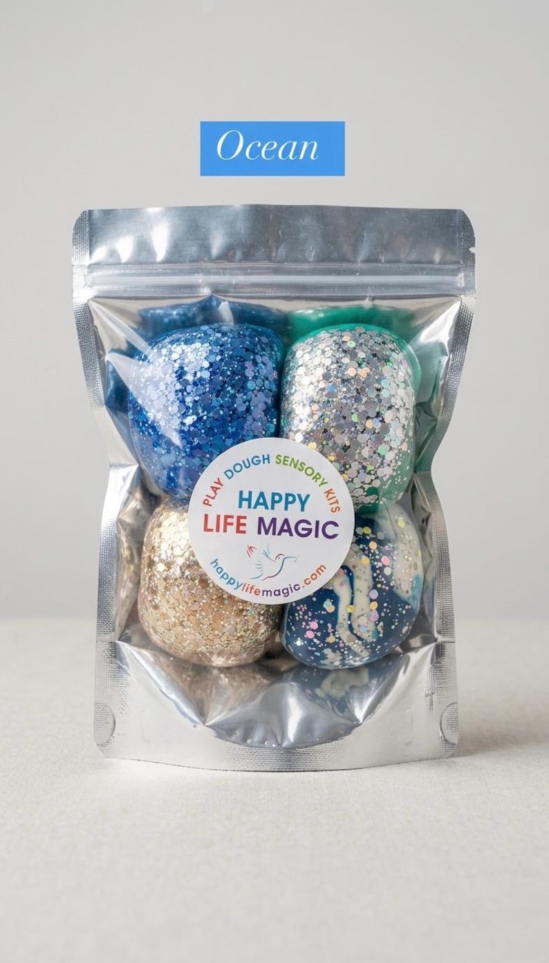 May include: A clear, resealable pouch containing four spheres of glittery play dough in shades of blue, silver, gold, and a marbled navy and white. A circular label on the front reads: Play Dough Sensory Kits, Happy Life Magic, happylifemagic.com.