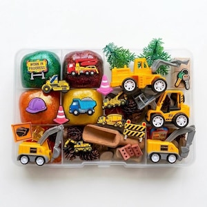 May include: A clear plastic divided storage container filled with sensory play items including green, red, and orange glittery dough, miniature yellow construction vehicles, small traffic cones, pinecones, a wooden scoop, and various construction-themed charms.