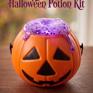 May include: An orange plastic jack-o'-lantern bucket with black facial features, filled with a thick, purple, glittery slime that drips over the rim. The text "Halloween Potion Kit" is printed in a dark purple font above the bucket.