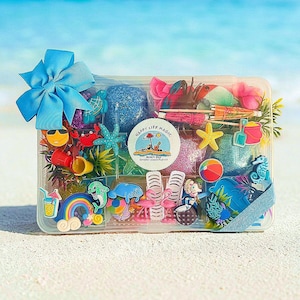 Pode incluir: Um recipiente de armazenamento retangular de plástico transparente preenchido com massa sensorial colorida e vários berloques de plástico com tema de praia, incluindo um caranguejo, estrela-do-mar, golfinho e flamingo. Um laço azul está preso.
