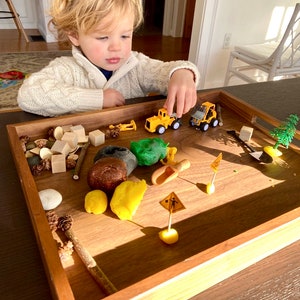 May include: A rectangular wooden tray containing a construction-themed sensory play set. The tray holds yellow plastic construction vehicles, small wooden blocks, brown and green modelling clay, miniature road signs, a small shovel, and pinecones.