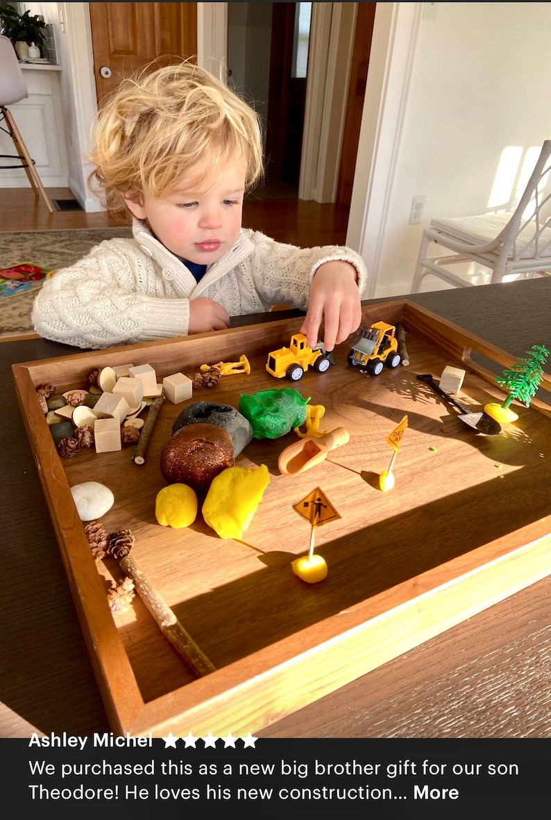 May include: A rectangular wooden tray containing a construction-themed sensory play set. The tray holds yellow plastic construction vehicles, small wooden blocks, brown and green modelling clay, miniature road signs, a small shovel, and pinecones.