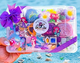 May include: A clear plastic box filled with mermaid-themed items, including figurines, decorative stones, and floral accents. The box is tied with a purple ribbon and bow. The text "Happy Life Magic" is on a circular sticker.