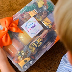May include: A clear plastic divided storage container filled with sensory play items including yellow construction vehicle toys, pinecones, and coloured dough. A bright orange ribbon is tied to the side, and a white label reads HAPPY LIFE MAGIC.