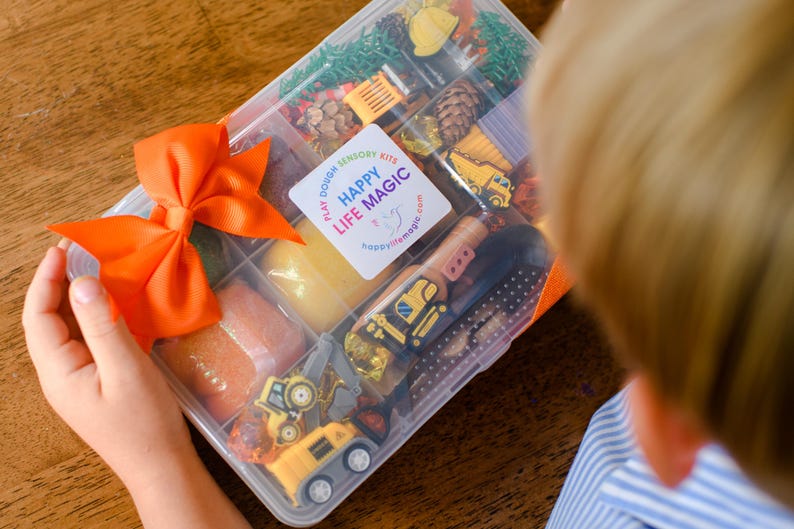 May include: A clear plastic divided storage container filled with sensory play items including yellow construction vehicle toys, pinecones, and coloured dough. A bright orange ribbon is tied to the side, and a white label reads HAPPY LIFE MAGIC.