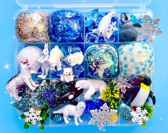 May include: A clear plastic storage container filled with various sensory play items including four textured slime balls, miniature plastic animal figures such as polar bears, a penguin, a seal, and a husky, plus snowflake ornaments and green foliage.