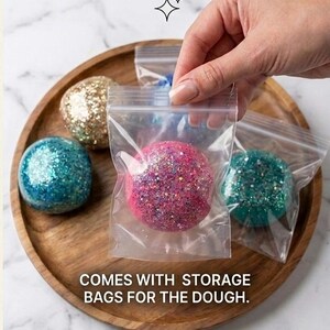 May include: Several spherical balls of glittery dough in various colours including pink, blue, gold, and teal. Each ball is individually packaged in a clear plastic resealable storage bag. The text on the image reads, COMES WITH STORAGE BAGS FOR THE DOUGH.
