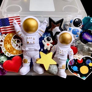 May include: A clear plastic storage box filled with space-themed items including two white astronaut figurines with gold helmets, a miniature American flag, a gold star, a moon-shaped cutout, a heart-shaped planet print, and various colorful beads.