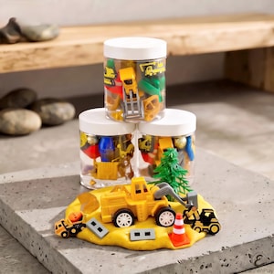 May include: Three clear plastic jars with white lids filled with small construction-themed toys, including yellow trucks and green trees. In front, a mound of yellow modeling clay features a yellow loader, a black excavator, and a traffic cone.
