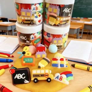 May include: Four clear plastic jars filled with colourful school-themed silicone charms, including yellow school buses, black chalkboards with ABC text, pencils, and math signs. Loose charms are arranged on a yellow dough base with crayons.