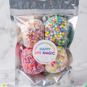 May include: A clear, resealable pouch containing four round balls of play dough in white, teal, pink, and off-white, each decorated with colourful sprinkles. A circular label reads "PLAY DOUGH SENSORY KITS HAPPY LIFE MAGIC" with a bird icon.