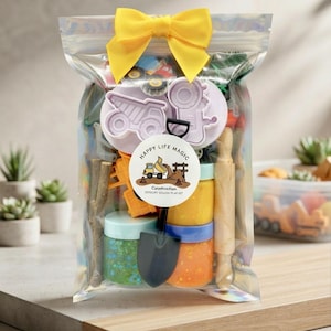 May include: A clear plastic pouch with a yellow bow containing a construction sensory dough play kit. Inside are small jars of colored dough, a wooden rolling pin, a black plastic shovel, and light purple construction vehicle cookie cutters.