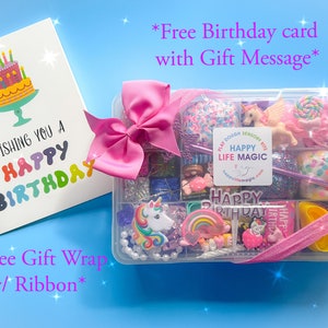 May include: A clear plastic storage box filled with colorful sensory items including unicorn figures, rainbow charms, and glittery dough, topped with a pink ribbon. Beside it is a white birthday card with a cake illustration and text.
