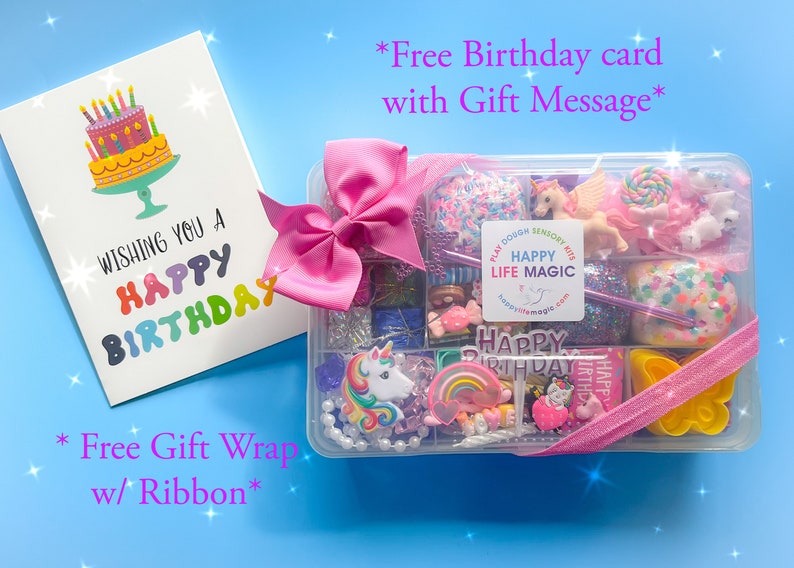 May include: A clear plastic storage box filled with colorful sensory items including unicorn figures, rainbow charms, and glittery dough, topped with a pink ribbon. Beside it is a white birthday card with a cake illustration and text.