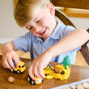 May include: A yellow toy dump truck and a yellow toy construction vehicle are positioned on a wooden surface next to a mound of yellow modelling clay, a small green plastic tree, and two small brown pinecones, with a storage container nearby.