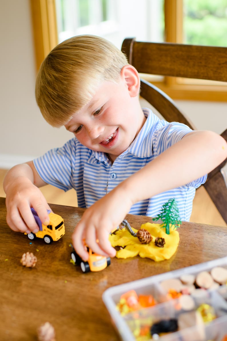 May include: A yellow toy dump truck and a yellow toy construction vehicle are positioned on a wooden surface next to a mound of yellow modelling clay, a small green plastic tree, and two small brown pinecones, with a storage container nearby.
