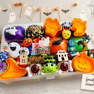 May include: A Halloween sensory kit in a clear plastic divided tray containing purple and orange slime, orange pumpkin and witch hat cookie cutters, plastic ghosts, eyeballs, a Frankenstein figure, a small M&M package, and wooden house cutouts.