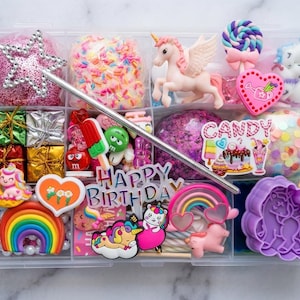 May include: A clear plastic organizer box filled with colorful craft supplies, including a silver star wand, a pink unicorn, a purple unicorn cookie cutter, rainbow charms, small gift boxes, and a sign that reads Happy Birthday and Candy.