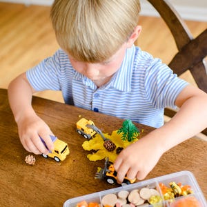 May include: A yellow toy dump truck and a yellow toy front loader are positioned on a wooden surface with yellow modelling dough, a small plastic pine tree, and pinecones. A clear plastic organiser box contains various craft supplies.
