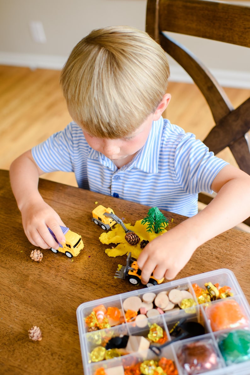 May include: A yellow toy dump truck and a yellow toy front loader are positioned on a wooden surface with yellow modelling dough, a small plastic pine tree, and pinecones. A clear plastic organiser box contains various craft supplies.