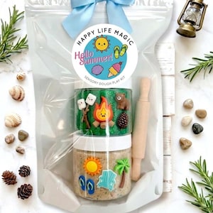 Op de afbeelding: Zomer Sensory Jar Bundle met twee potten sensorische deeg, één met een kampeerthema en de andere met een strandthema. Inclusief een deegroller. Het pakket is vastgebonden met een lichtblauwe strik. De tekst "Hello Summer!" staat op het label.