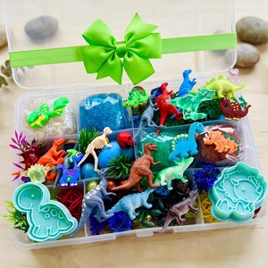 May include: A clear plastic divided storage container filled with various small dinosaur figurines, colorful slime, and two light blue dinosaur-shaped cookie cutters. The lid is decorated with a bright lime green ribbon tied in a bow.