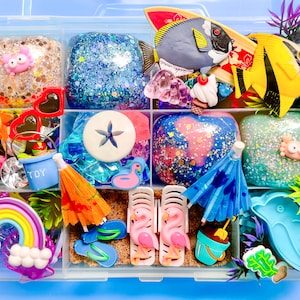 May include: A clear plastic storage box filled with colorful summer-themed slime and toys. The box has nine compartments, each filled with a different type of slime or toy. Some of the toys include a rainbow, a starfish, a sun, a dolphin, a flamingo, and an umbrella. The slime is sparkly and glittery.