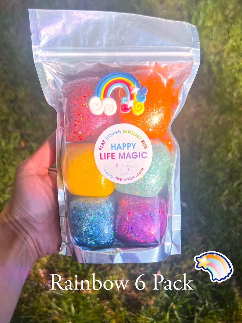May include: A clear plastic pouch containing six individual portions of colourful play dough in red, orange, yellow, green, blue, and purple, each with glitter. A rainbow charm sits on top, and a circular label reads PLAY DOUGH SENSORY KITS HAPPY LIFE MAGIC.