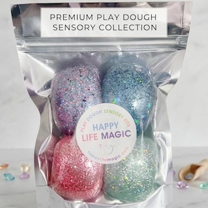 May include: A clear, resealable pouch containing four rounded portions of glittery play dough in pink, blue, purple, and green. The label reads PREMIUM PLAY DOUGH SENSORY COLLECTION, Unicorn, and HAPPY LIFE MAGIC with a small bird logo and website.
