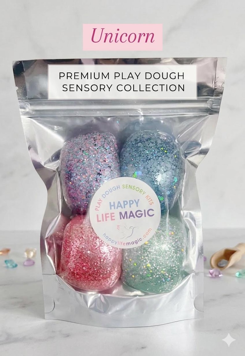 May include: A clear, resealable pouch containing four rounded portions of glittery play dough in pink, blue, purple, and green. The label reads PREMIUM PLAY DOUGH SENSORY COLLECTION, Unicorn, and HAPPY LIFE MAGIC with a small bird logo and website.