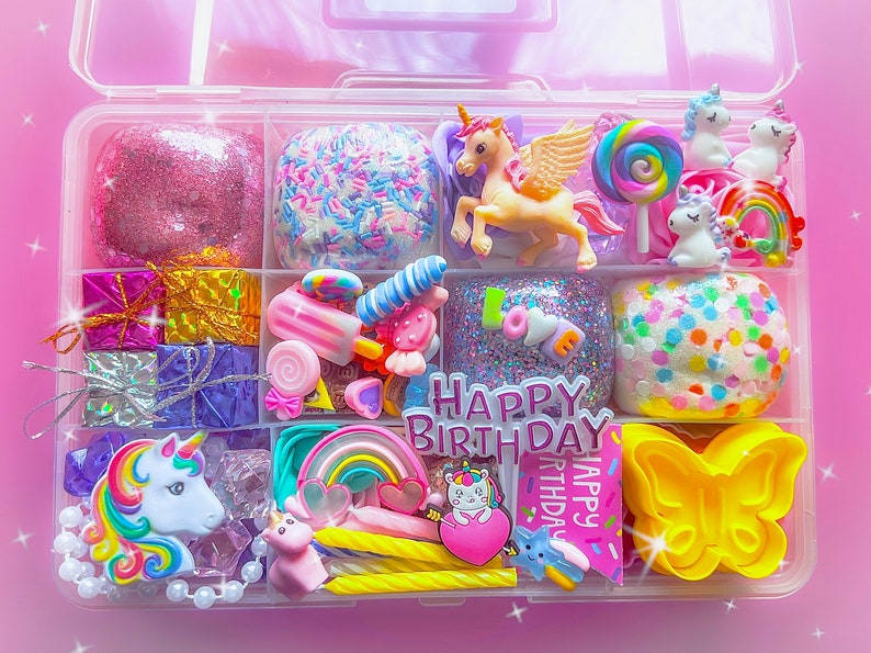 May include: A clear plastic organizer box filled with colorful craft supplies including pink glitter slime, a winged unicorn figurine, rainbow charms, small gift boxes, a yellow butterfly mold, and a sign that reads HAPPY BIRTHDAY.