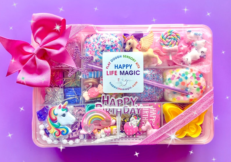 May include: A rectangular pink plastic storage container filled with various colorful sensory play items, including a unicorn figurine, a rainbow charm, a yellow butterfly mold, and a Happy Birthday sign. A large pink bow is attached to the top left.