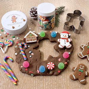 May include: A gingerbread-themed playdough kit featuring a brown dough wreath decorated with miniature candy-shaped charms, a snowman, and a house. Includes a metal gingerbread man cutter, a jar of colorful charms, and a white tin labeled Happy Life Magic.