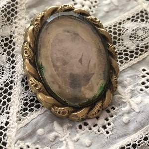 May include: A gold-toned oval locket with a glass front and a braided design around the edge. The locket is empty and has a faded image inside.