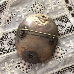 May include: A silver oval brooch with a pin on the back. The brooch has a textured surface and a worn look.