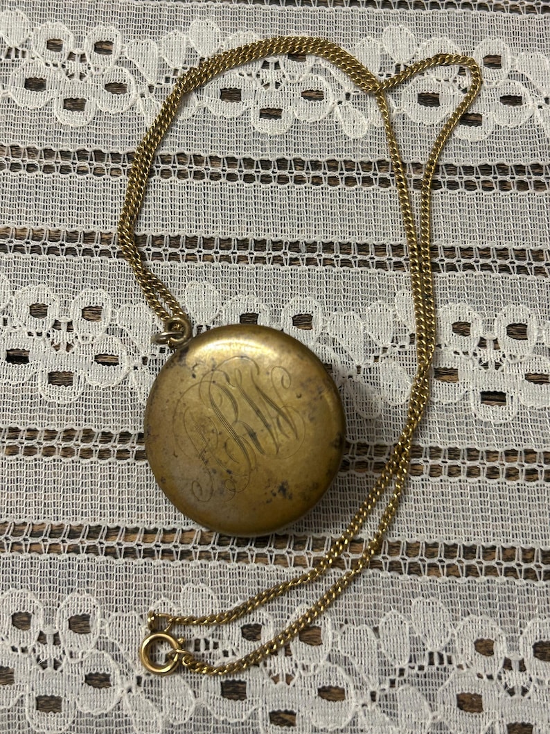 Puede incluir: Un collar de cadena dorada con un colgante redondo en forma de medall&oacute;n. El medall&oacute;n tiene un monograma grabado.