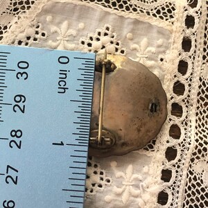 May include: A silver-toned brooch with a detailed design, measuring approximately 1 inch in diameter. The brooch has a pin and catch closure.