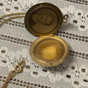 Puede incluir: Un collar de oro con un medall&oacute;n y una cadena. El medall&oacute;n est&aacute; abierto y revela una foto de un hombre en su interior. El medall&oacute;n est&aacute; sobre una superficie de encaje blanco.