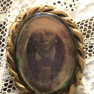 May include: A gold-toned oval locket with a braided rope design around the edge. The locket contains a faded black and white photograph of a person.